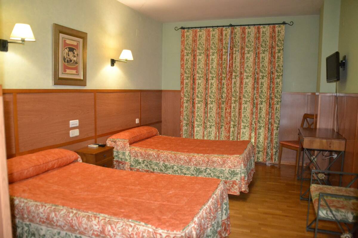 Quarto de hotel com duas camas, cabeceira de madeira, uma mesa pequena, uma cadeira, televisão e cortina com padrão floral.