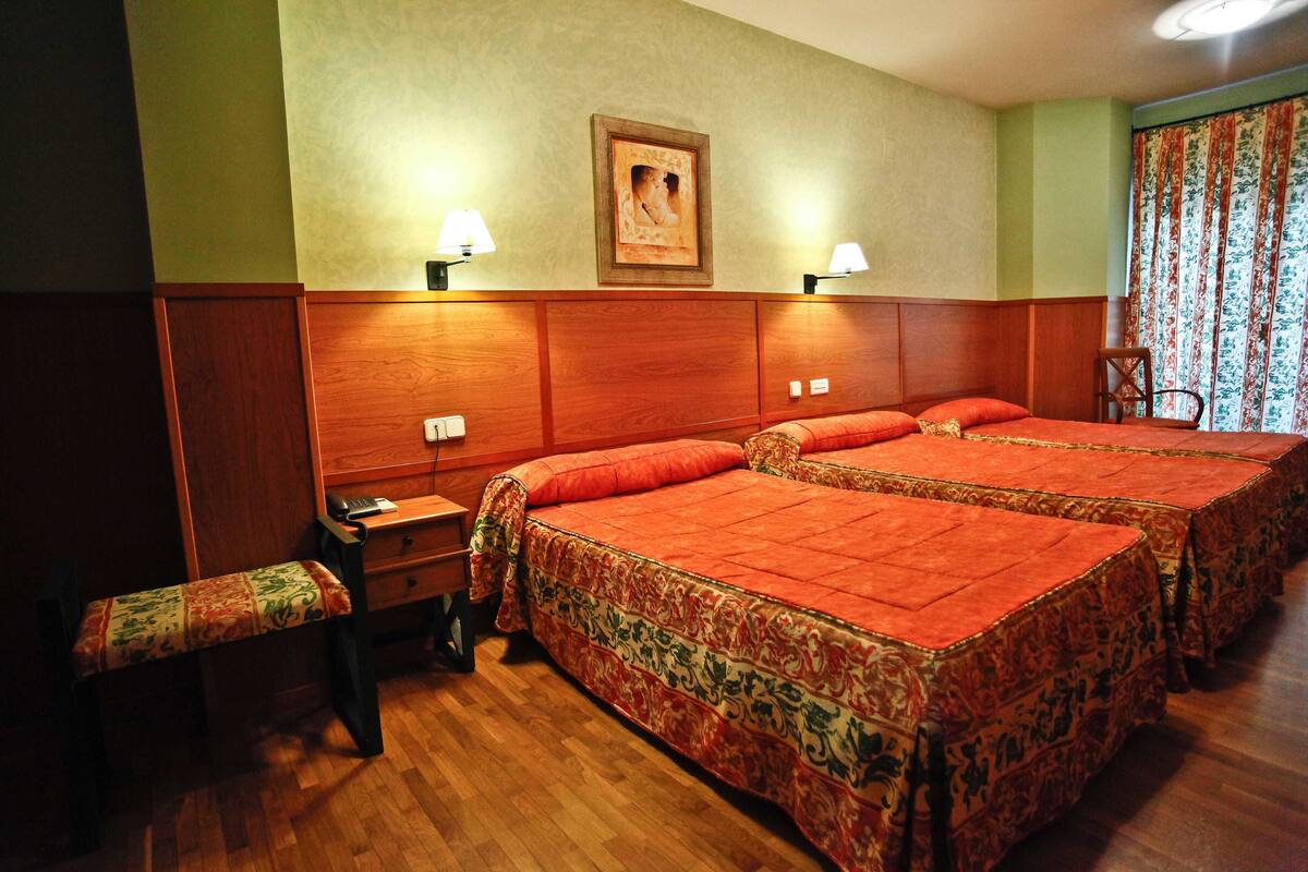 Quarto de hotel com duas camas, um banquinho, uma mesa de cabeceira e uma luminária fixada na parede.