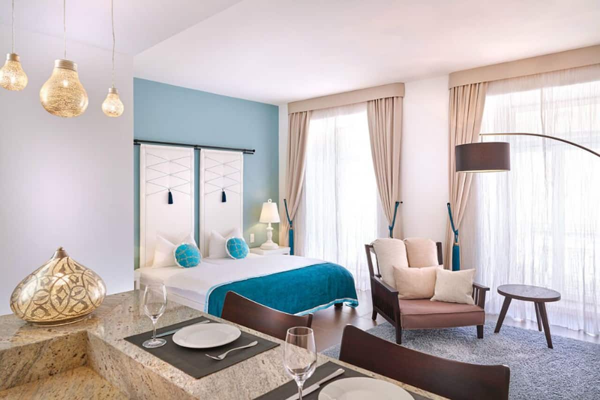 Een hotelkamer met een bed, een zithoek met een bank en fauteuils, een eettafel gedekt voor twee personen, en een groot raam met gordijnen.