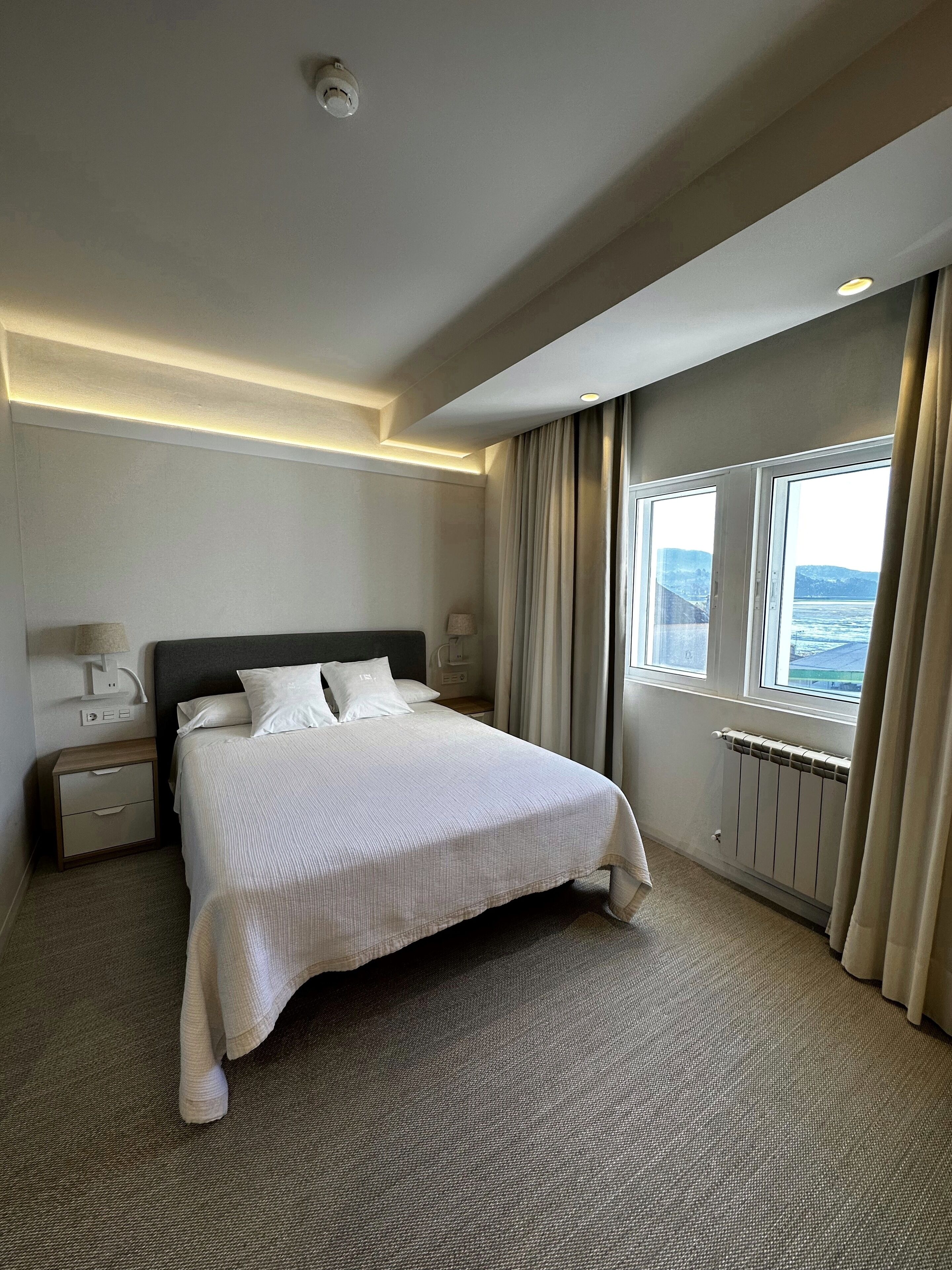 A hotel room with a large bed, bedside tables, a nightstand, a window with a view, and a ceiling light.