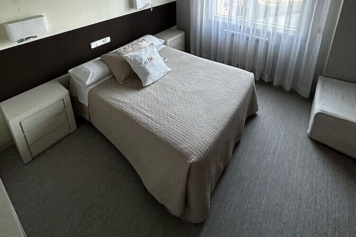 Una habitación de hotel con una cama grande, mesitas de noche, cabecera, una ventana con cortinas y una lámpara en el techo.