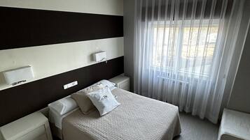 Una habitación de hotel con una cama grande, mesitas de noche, una lámpara en el techo y una ventana con cortinas translúcidas.