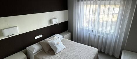 Una habitación de hotel con una cama grande, mesitas de noche, una lámpara en el techo y una ventana con cortinas translúcidas.
