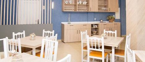 A dining area with white wooden tables and chairs, a kitchen with light wood cabinets, and a blue accent wall.