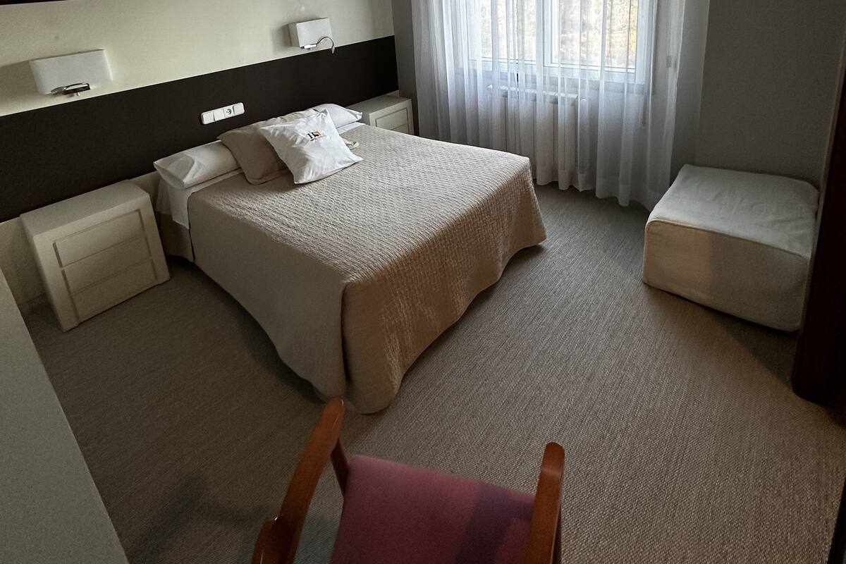 Una habitación de hotel moderna con una cama grande, una mesita de noche, una silla y un ventanal con cortinas.