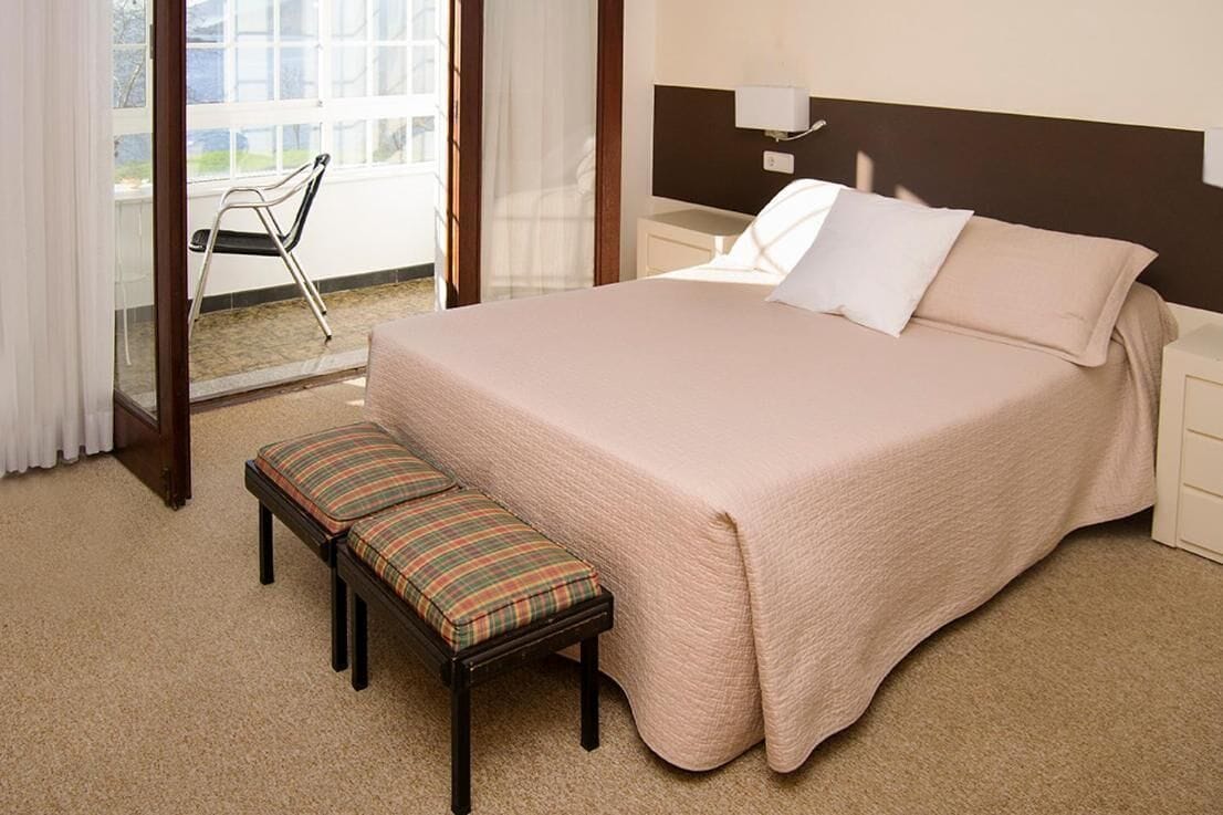 A hotel room with a large bed, a bench with a plaid cushion, a bedside table, and a window with a view of the outdoors.