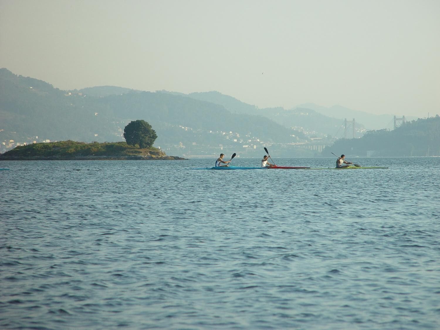 Three kayakers on a calm body of water with a distant shoreline and hills.