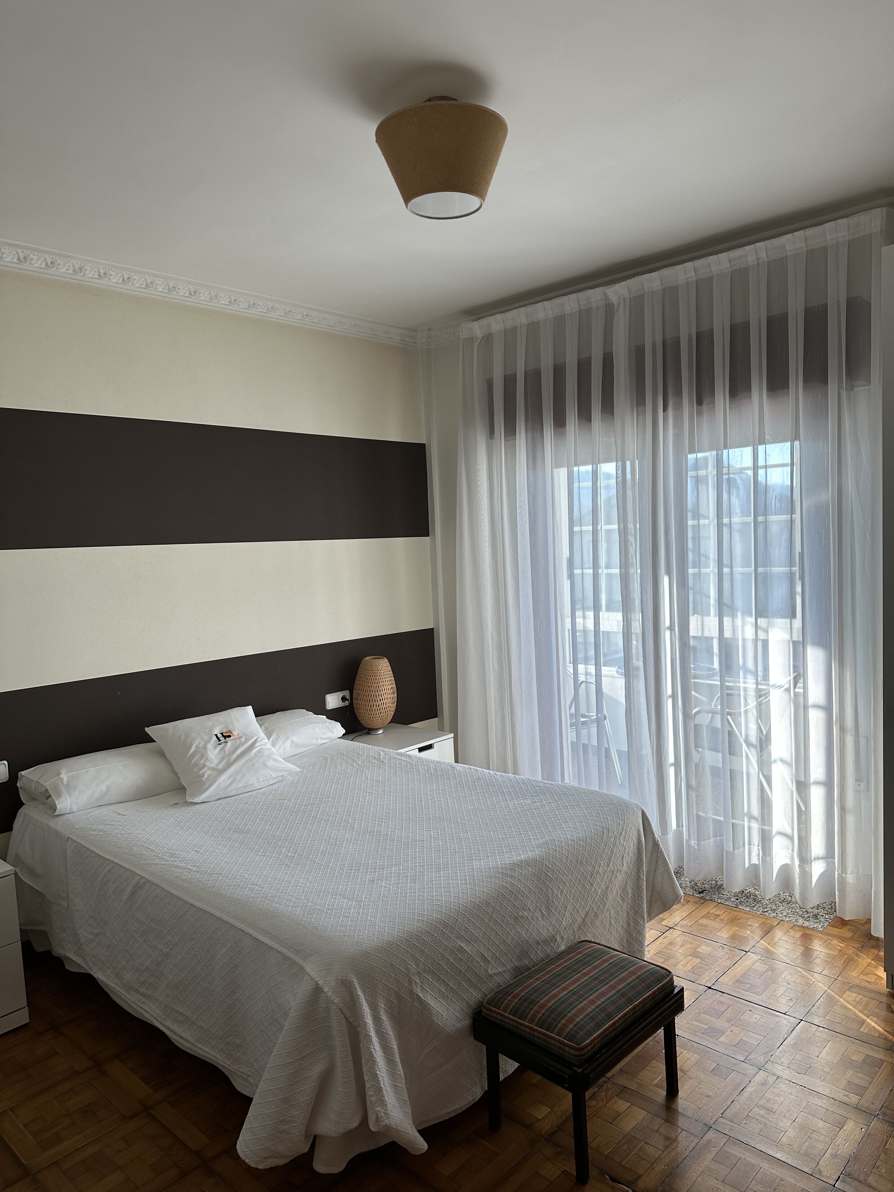 Cama bem arrumada com lençóis brancos, mesa de cabeceira com luminária e cortinas leves cobrindo a janela.