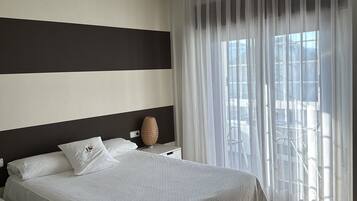 Cama bem arrumada com lençóis brancos, mesa de cabeceira com luminária e cortinas leves cobrindo a janela.