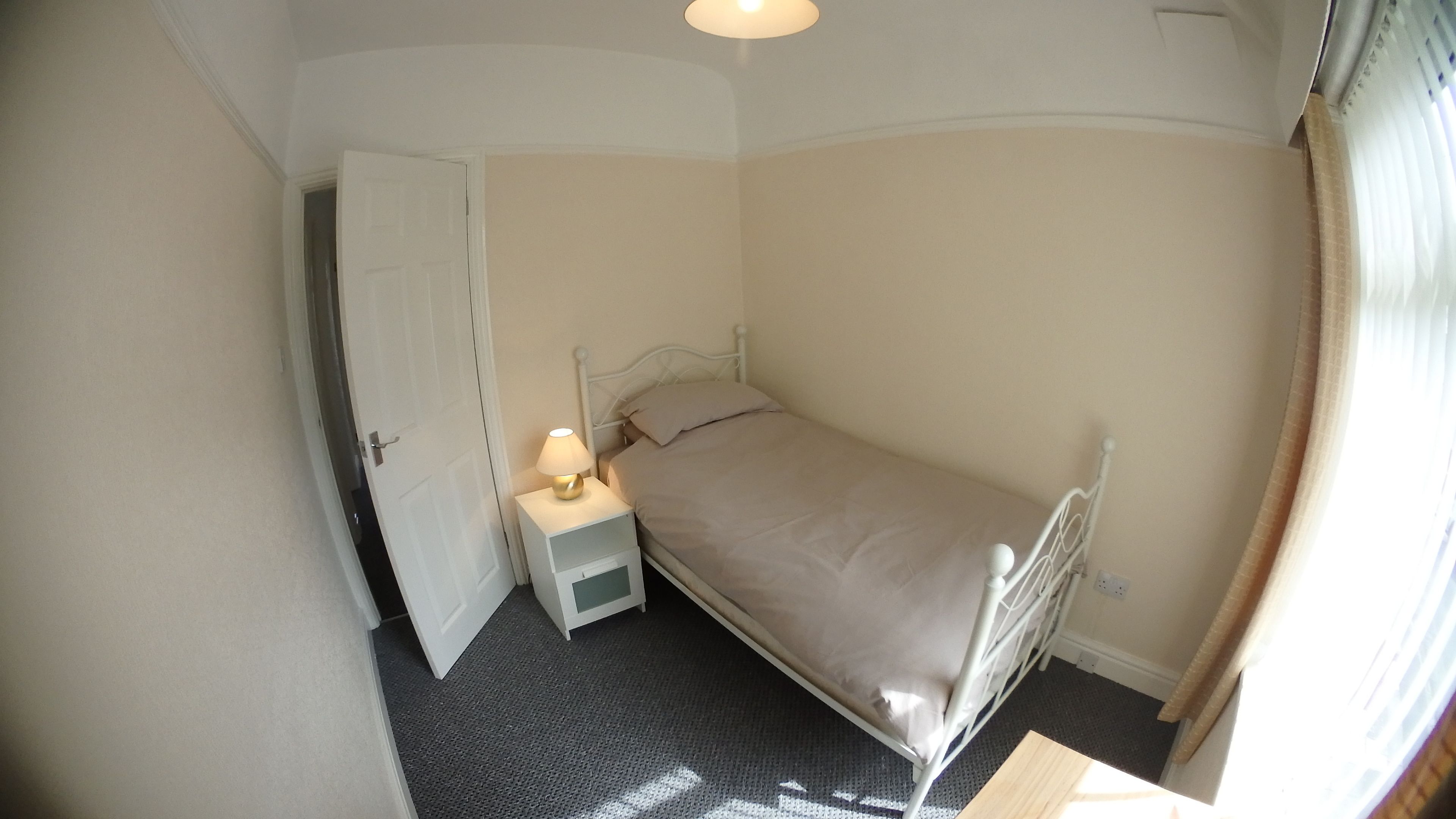 A small, single-bed room with a white metal bed frame, a bedside table with a lamp, and a door leading to another room.
