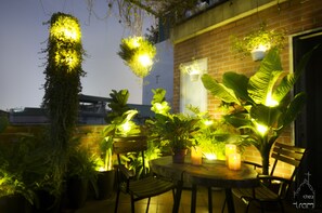 Eine Außenpatio-Terrasse mit rundem Tisch, Stühlen, Kübelpflanzen, hängenden Pflanzen und Wandleuchten.