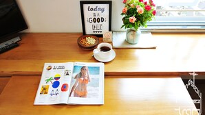 A wooden desk with a magazine, a framed quote, a bowl of nuts, a cup of coffee, and a vase of flowers.