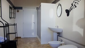 A bathroom with a white sink, toilet, and bidet. There is a mirror on the wall and a black metal shelf with a heart-shaped decoration. - MyRoom CenterStation (Bologna)