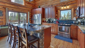 A spacious kitchen with wooden cabinets, a large island with bar stools, stainless steel appliances, and a view of the forest through large windows. - Access to Rocky Mtn Nat'l Park: Grand Lake Cabin (Grand Lake)
