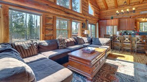 A log cabin interior with a large L-shaped sectional sofa, a wooden coffee table, and a kitchen area with wooden cabinets and a bar. - Access to Rocky Mtn Nat'l Park: Grand Lake Cabin (Grand Lake)