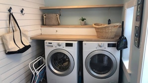 A compact laundry room with two front-loading washing machines, a wicker basket on a shelf, a small potted plant, and a wall-mounted drying rack. - Coastal Charmer, Fire Pit & Two Blocks to Beach (Cape Charles)