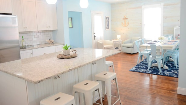 A modern kitchen with white cabinets, a granite countertop, and bar stools. There is a dining area with a table and chairs, and a blue rug on the floor. - Coastal Charmer, Fire Pit & Two Blocks to Beach (Cape Charles)