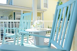 A porch with a round table and two blue chairs, a white vase with flowers, and a yellow building in the background. - Coastal Charmer, Fire Pit & Two Blocks to Beach (Cape Charles)