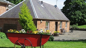 A stone building with a sloped roof, a satellite dish, and a wheelbarrow filled with flowers. - Harleyburn Cottages (Melrose)