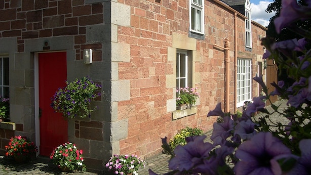 A stone building with a red door, windows with flower boxes, and a brick pathway. - Harleyburn Cottages (Melrose)