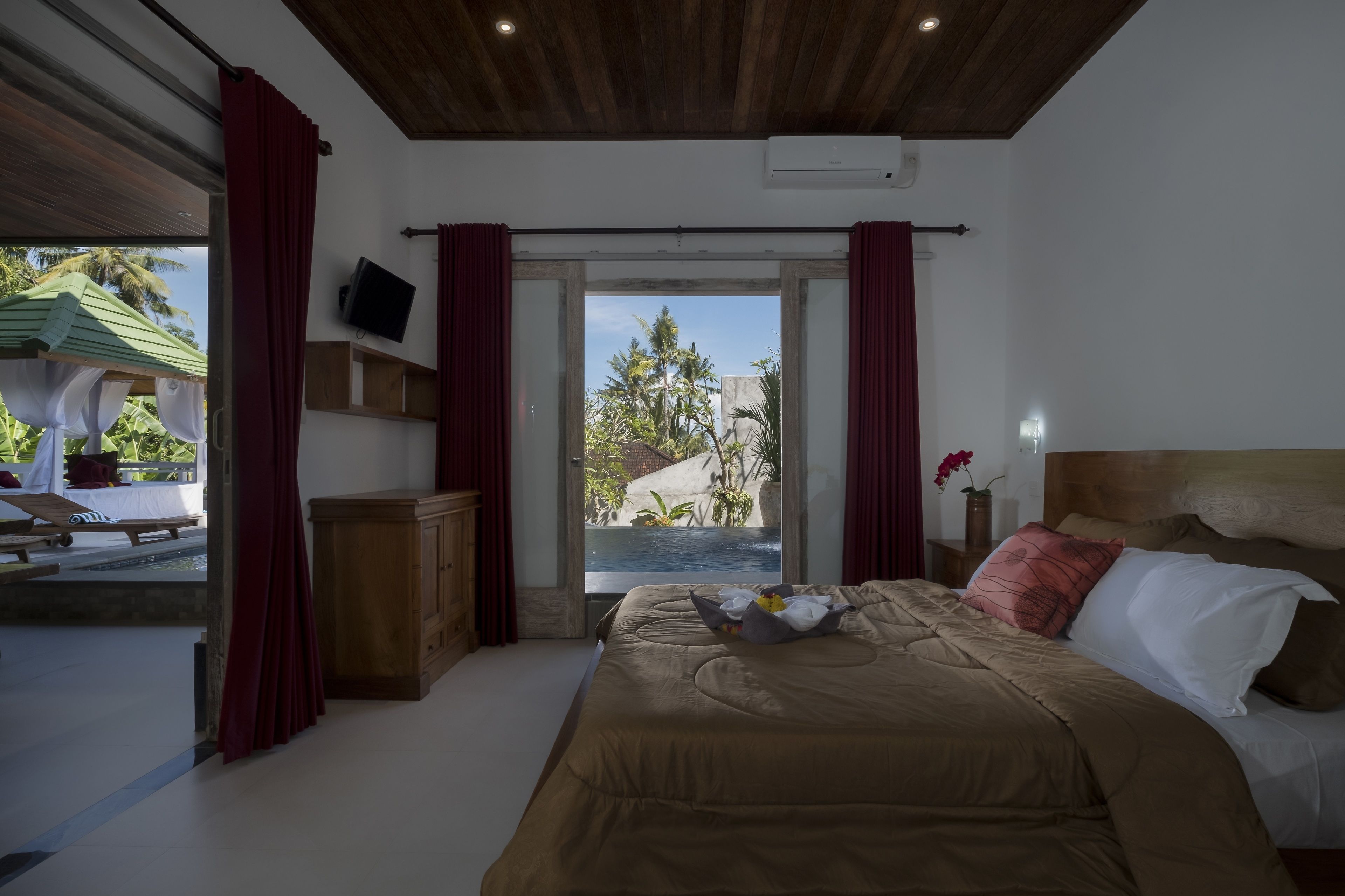 A bedroom with a bed, a television, a wooden headboard, a nightstand, and a view of a pool and palm trees.