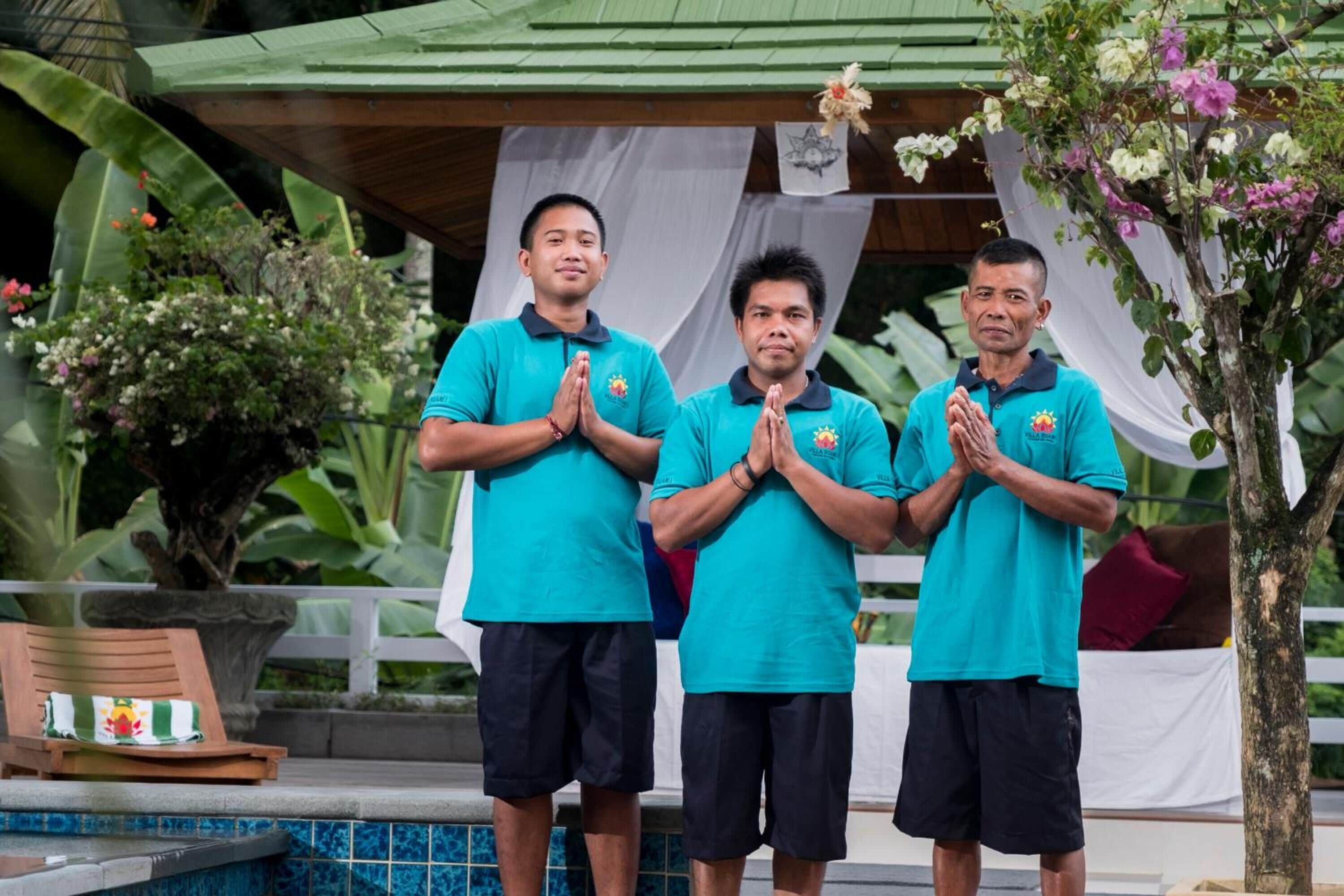 Trois personnes, vêtues de chemises turquoise, se tiennent près d’une piscine, avec un paysage tropical en arrière-plan.