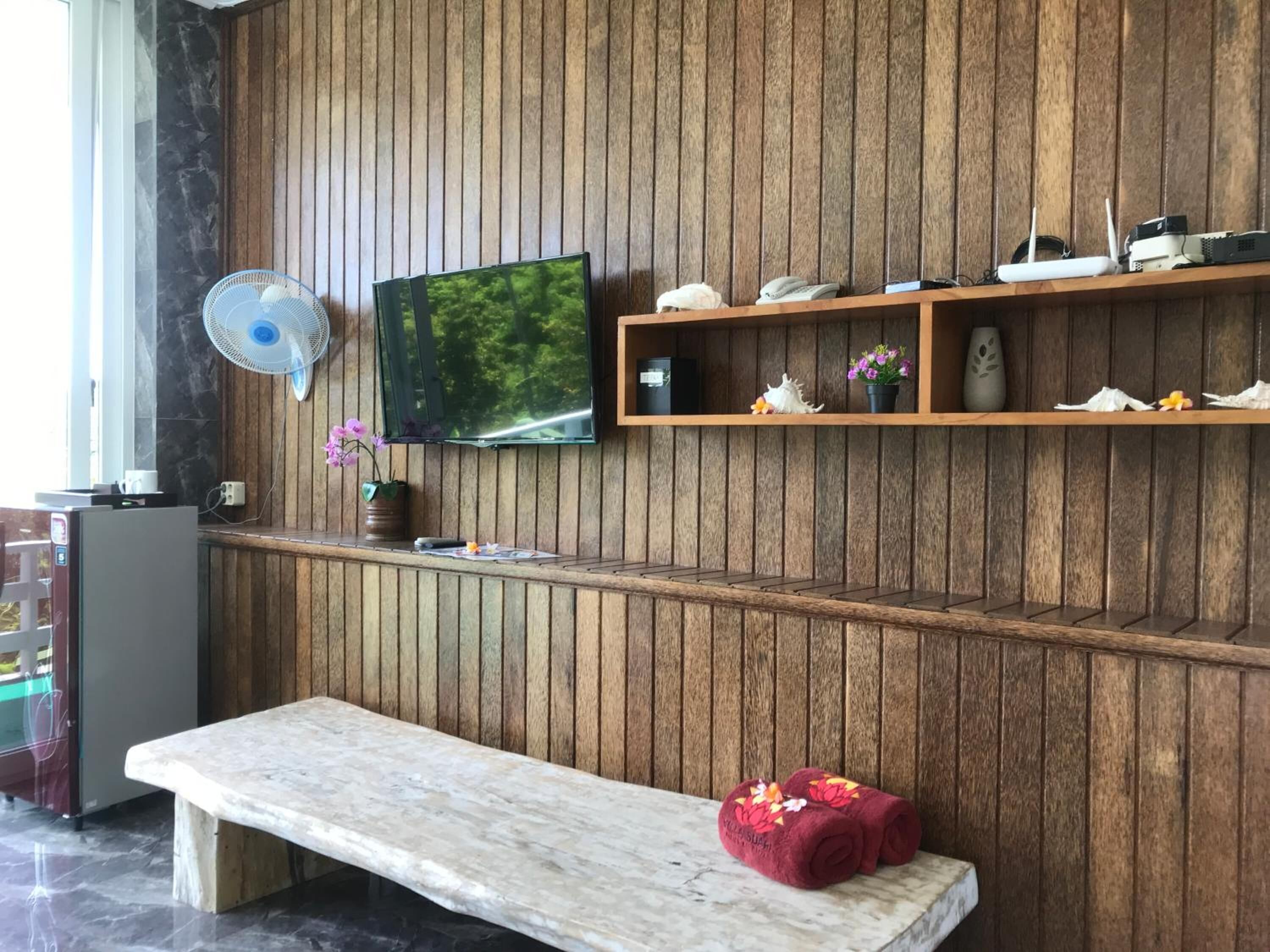 A room with a wooden panel wall, a flat-screen TV, a fan, a bench, and a shelf with various items.