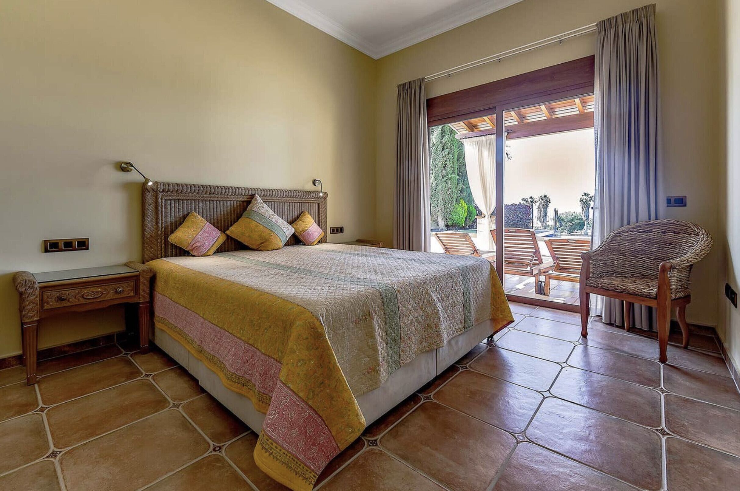 A bedroom with a bed, a nightstand, a wicker chair, and a view of the outdoors through a sliding door.