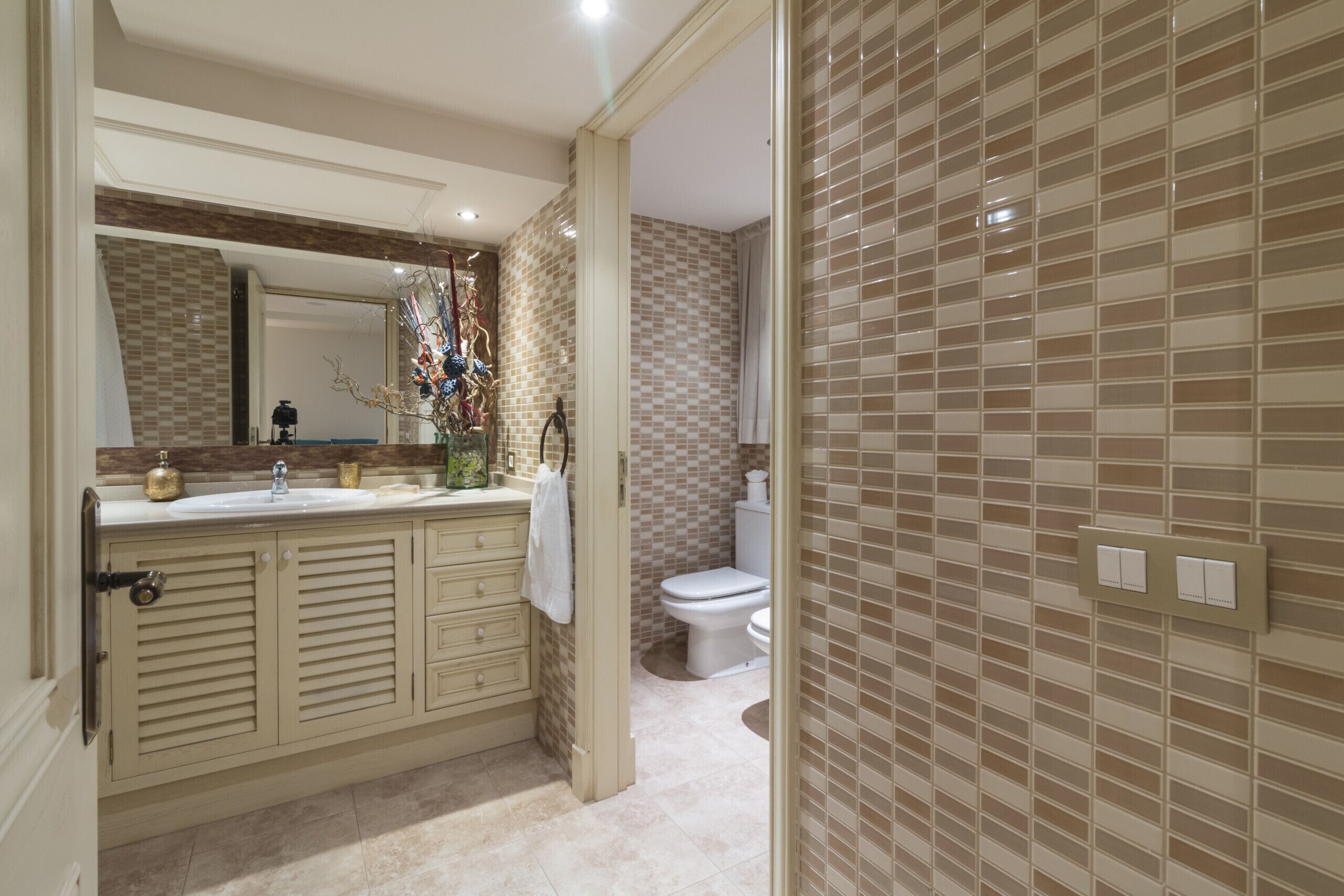 Een badkamer met een beige wastafelmeubel, een grote spiegel, een toilet en een betegelde muur.