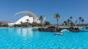 A large swimming pool with clear blue water, surrounded by palm trees and a distinctive white building with a curved roof.