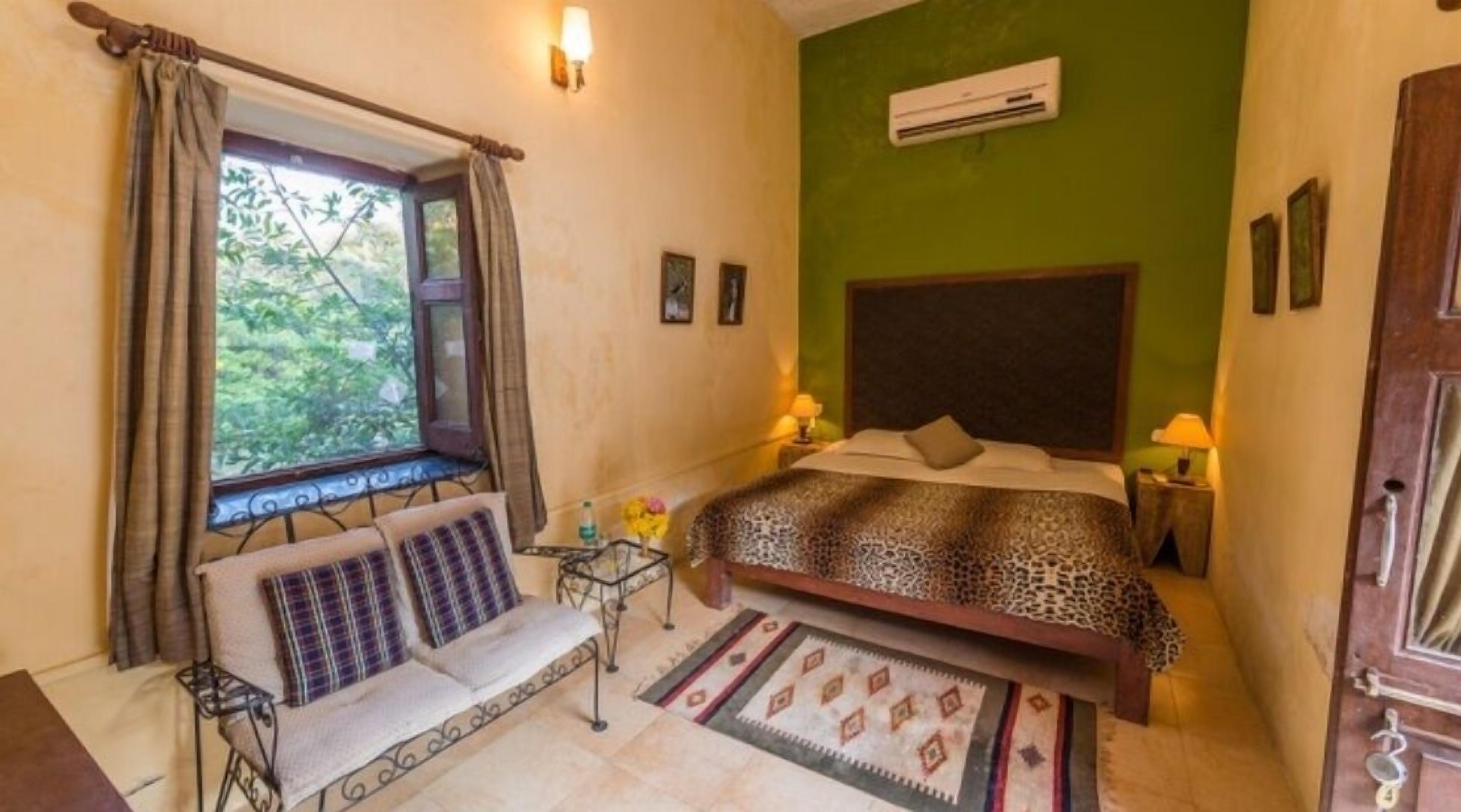 A bedroom with a bed, a sofa, a window with curtains, a bedside table, and a wall-mounted air conditioner.