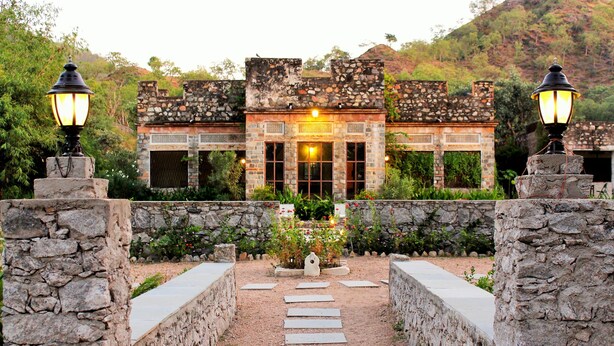 Un edificio de piedra con una entrada central, rodeado de un camino de piedra y delimitado por muros de piedra.