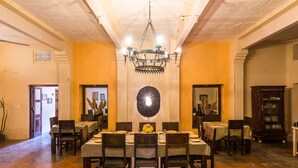 Comedor con una mesa grande dispuesta para seis personas, rodeada de sillas, con una araña colgante y un reloj en la pared.