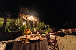 Un área de comedor al aire libre con una mesa dispuesta para dos personas, rodeada de muros de piedra y vegetación.