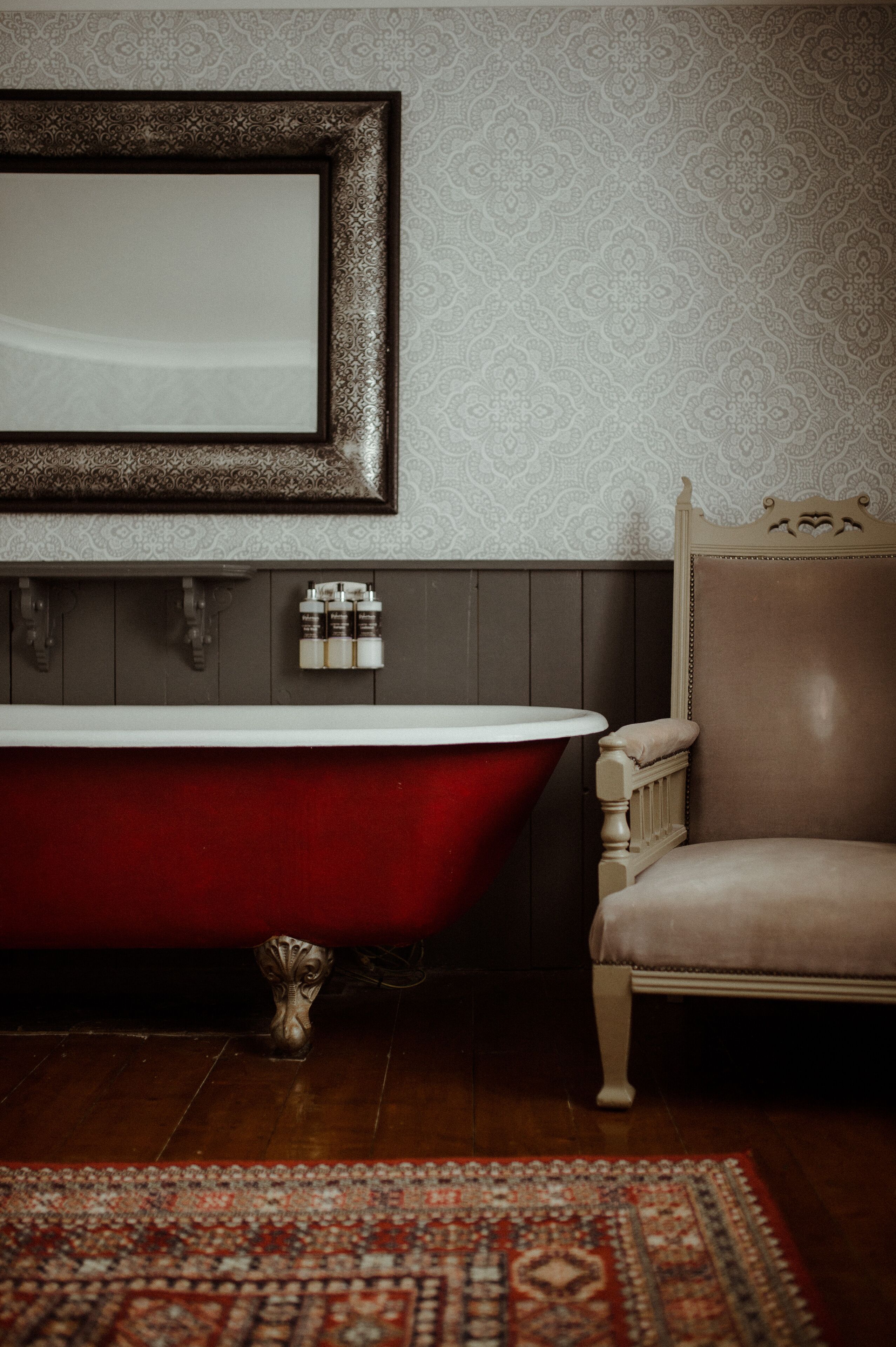 A bathroom with a red clawfoot bathtub, a wooden chair, a mirror with a decorative frame, and a patterned wall.