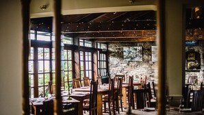 A restaurant with wooden tables and chairs, large windows, and a stone wall. - The George Hotel (Inveraray)