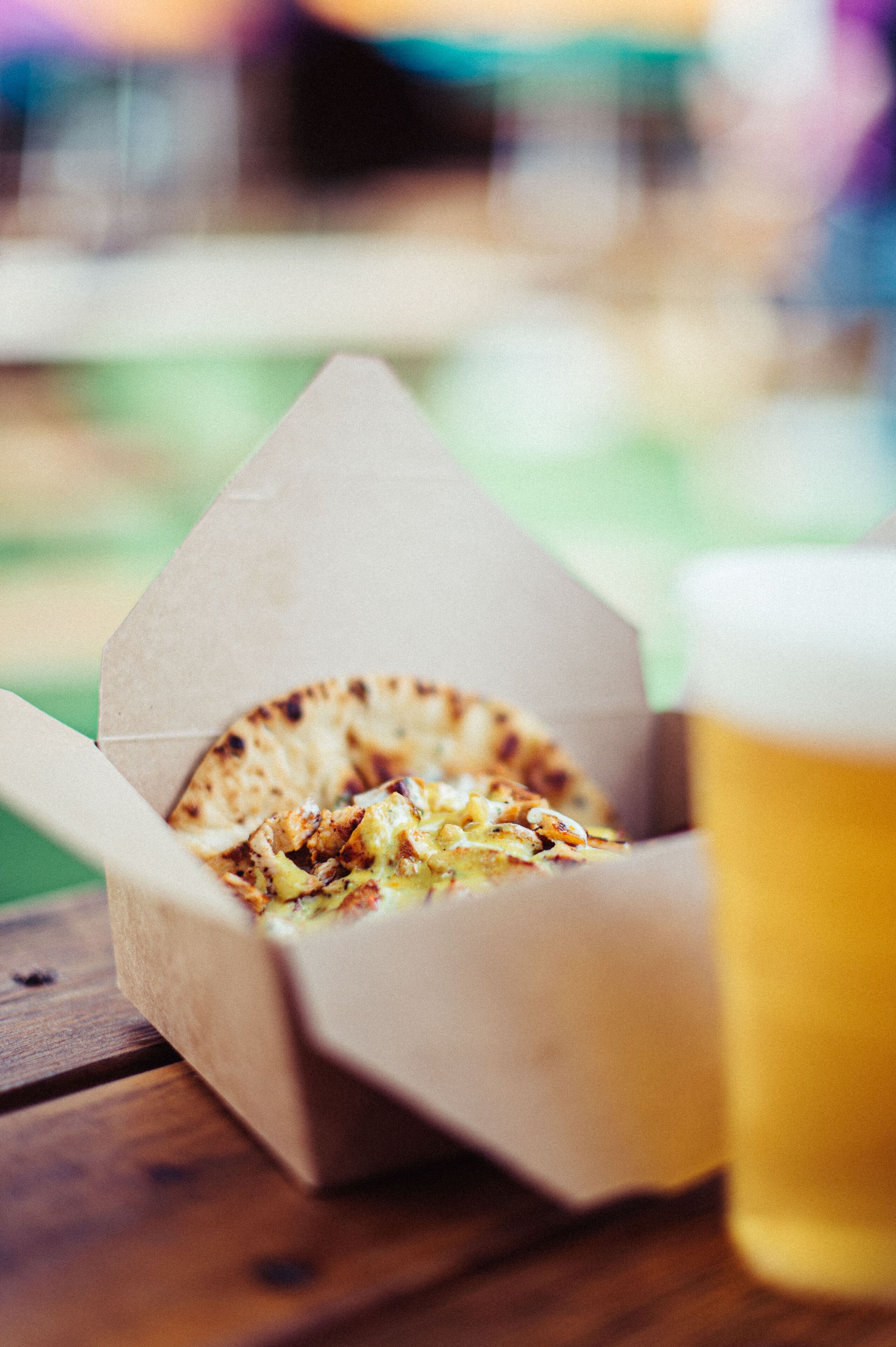 A paper food container with a food item and a glass of beer on a wooden surface.