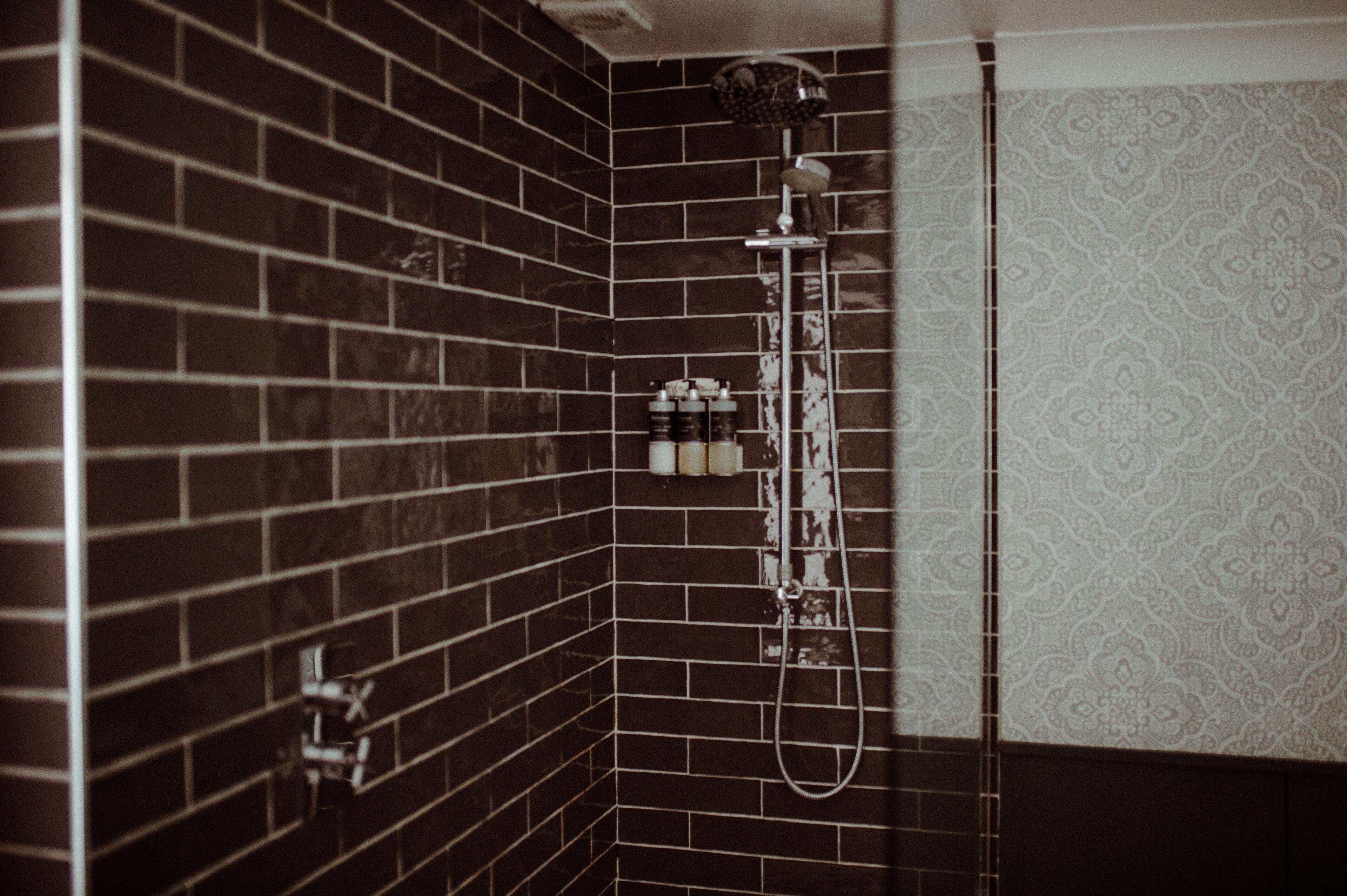 A bathroom with dark tiled walls, a shower with a handheld showerhead, and a patterned wall.