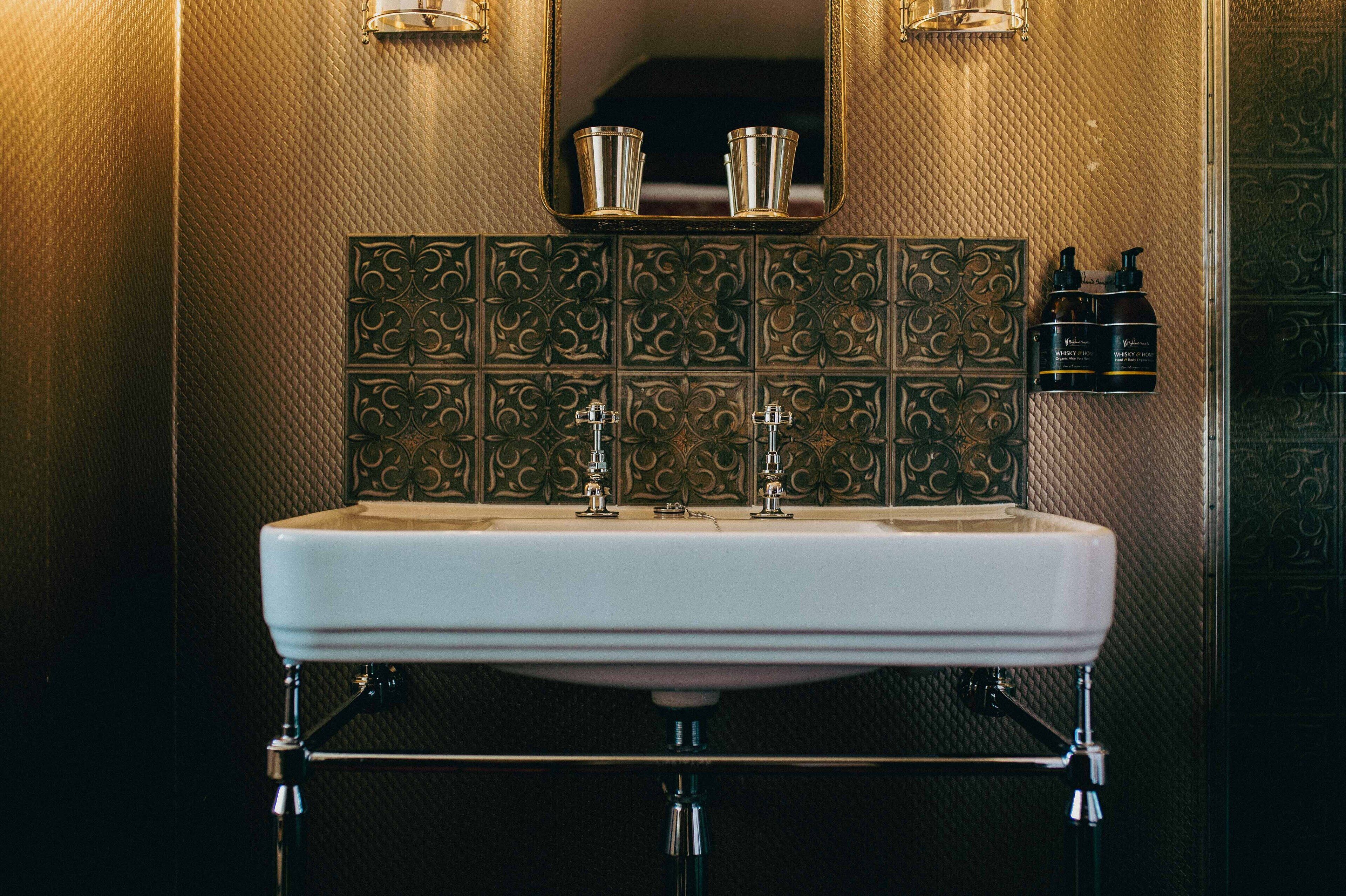 A bathroom with a white sink, two faucets, and a mirror above. There are two bottles of hand soap on the wall.