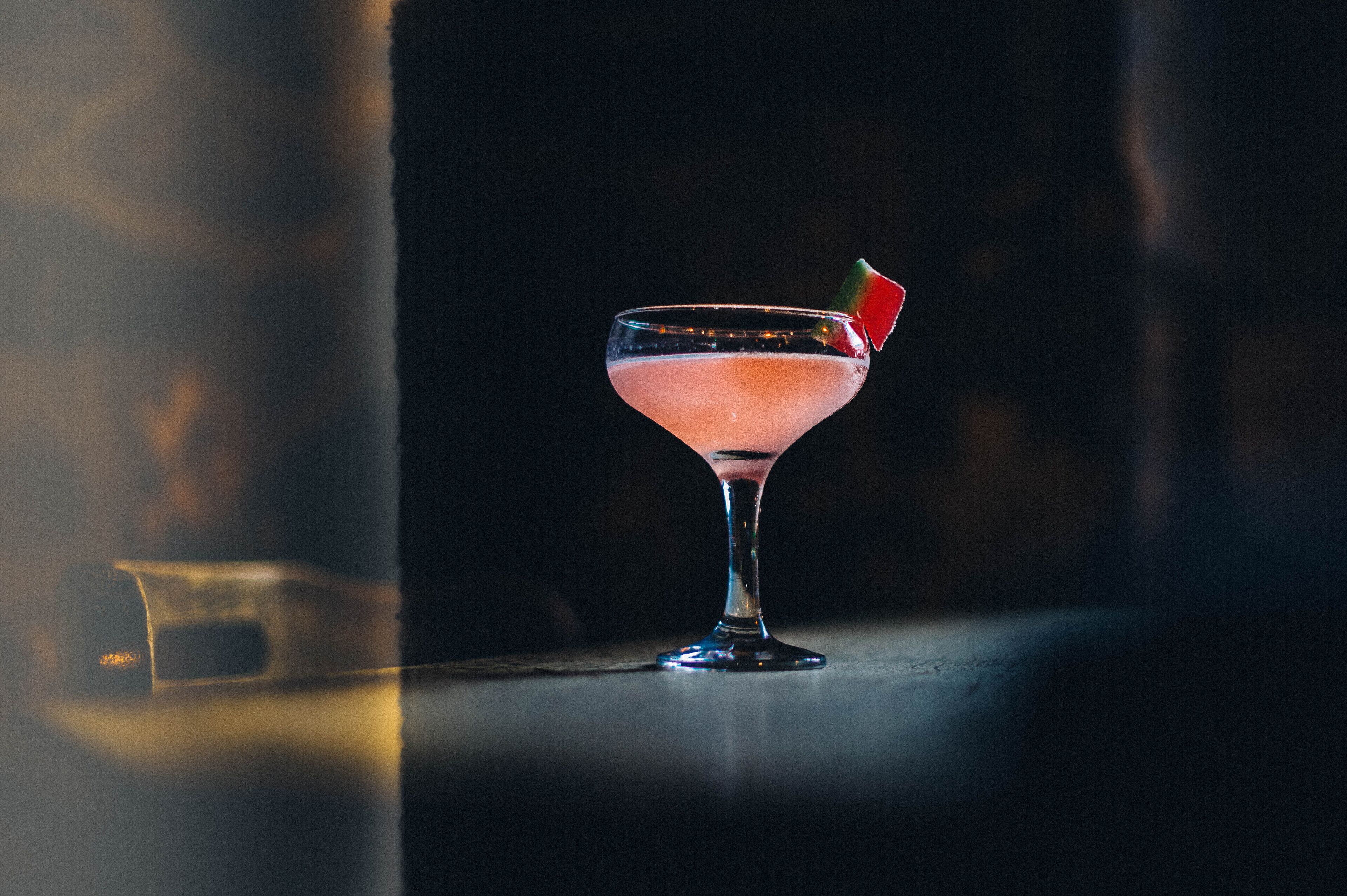 A cocktail in a martini glass with a garnish, placed on a reflective surface.