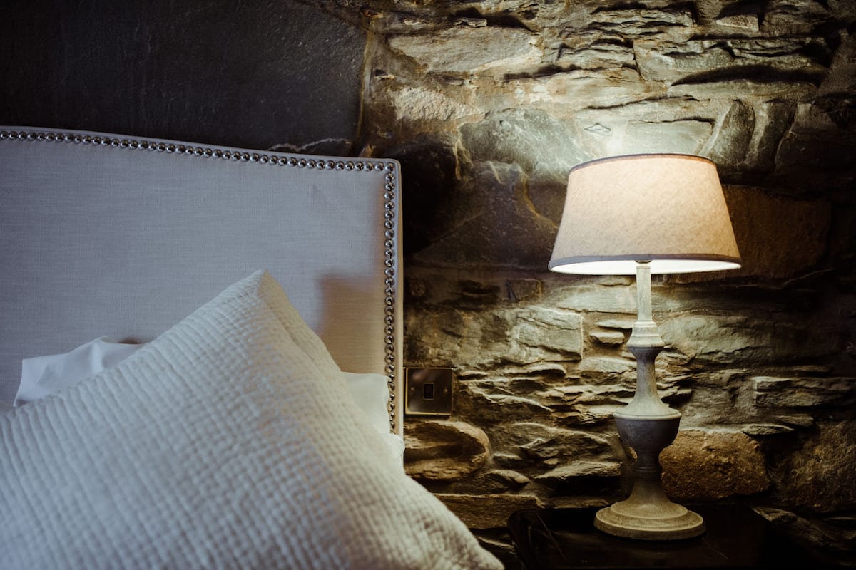 A bed with a headboard and a lamp against a stone wall.