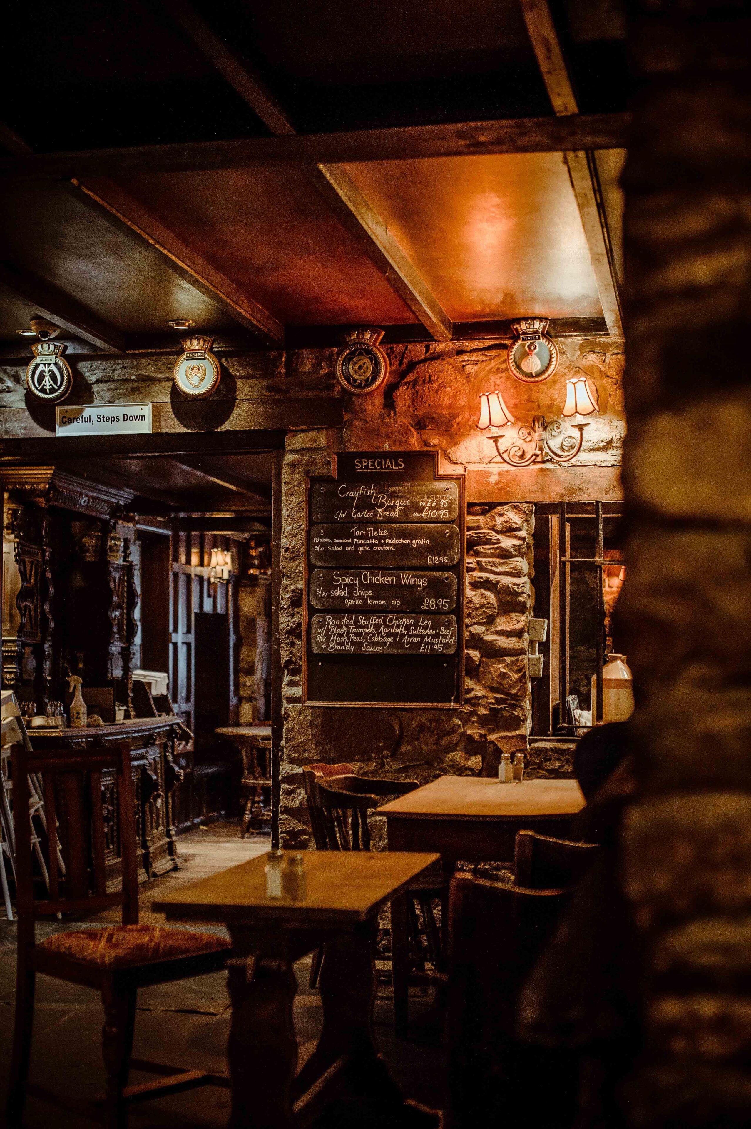 A cozy restaurant interior with wooden tables and chairs, a stone wall, and a menu board listing dishes.