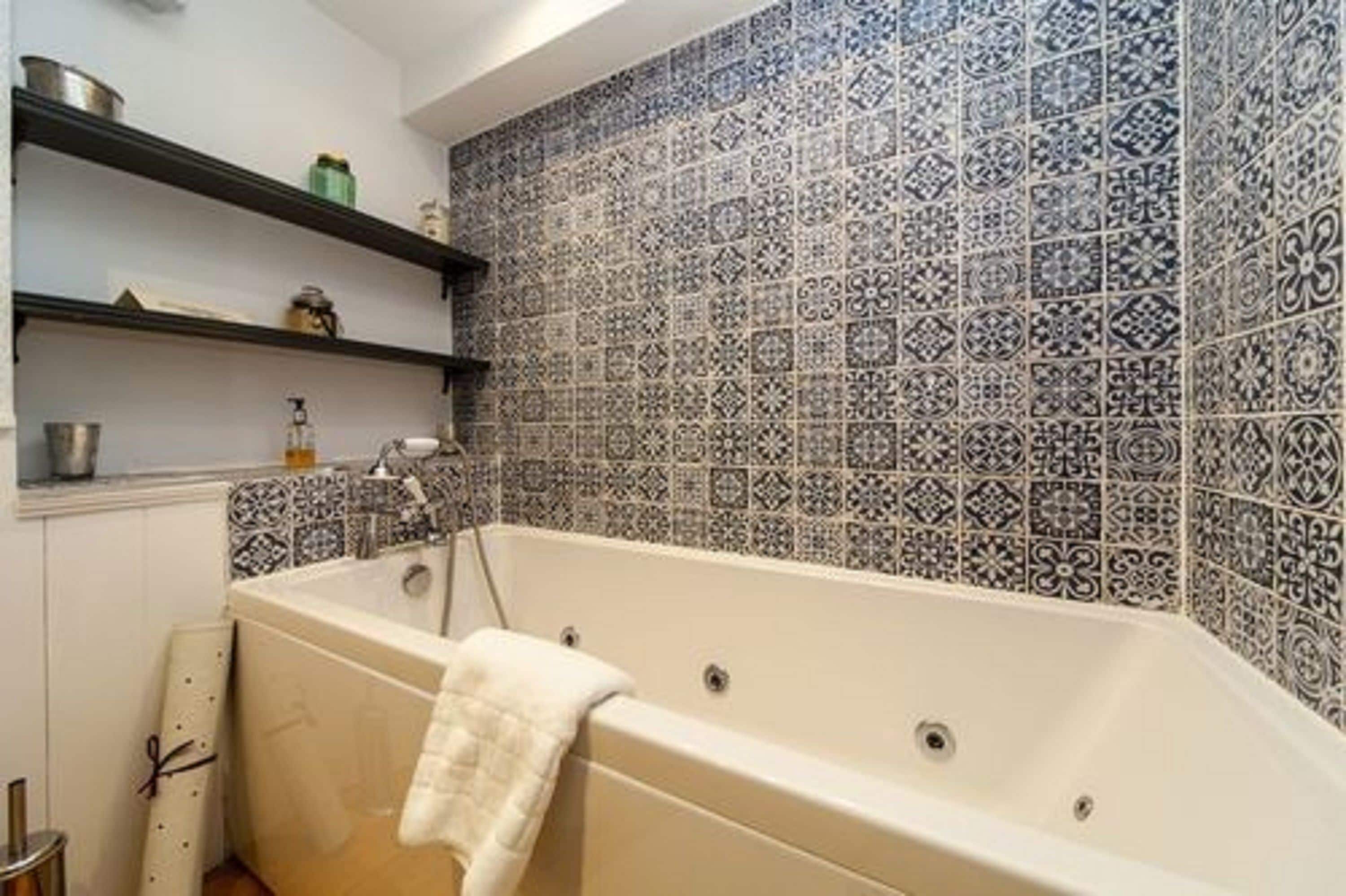 Um banheiro com banheira, prateleiras com produtos de higiene e azulejos estampados.