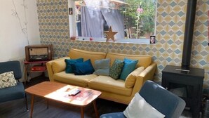 A living room with a yellow sofa, blue armchairs, a wooden coffee table, and a black wood stove. - House with garden in the city center (Marseille)