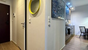 Un interior moderno con suelo de madera, una puerta con espejo, un cuadro enmarcado y una zona de cocina con armarios y una silla.