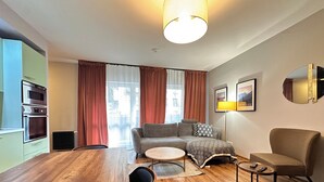 Una sala moderna con una mesa de comedor de madera, un sofá, una mesa de centro y una lámpara. Hay una zona de cocina con horno y radiador.