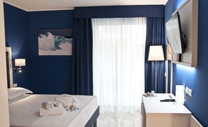 Habitació d'hotel amb llit, taula de nit, làmpada i una finestra gran amb cortines.