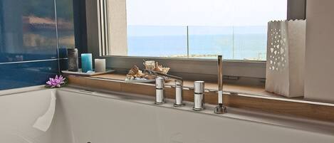 A bathroom with a bathtub, a window with a view of the sea, and a shelf with decorative items.