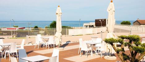 A terrace with white chairs and tables, a tree, and a view of the sea.