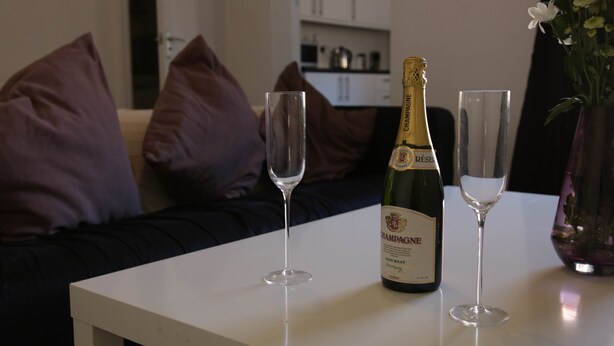 Una bottiglia di champagne e due bicchieri su un tavolo bianco in un soggiorno con un divano e dei cuscini.
